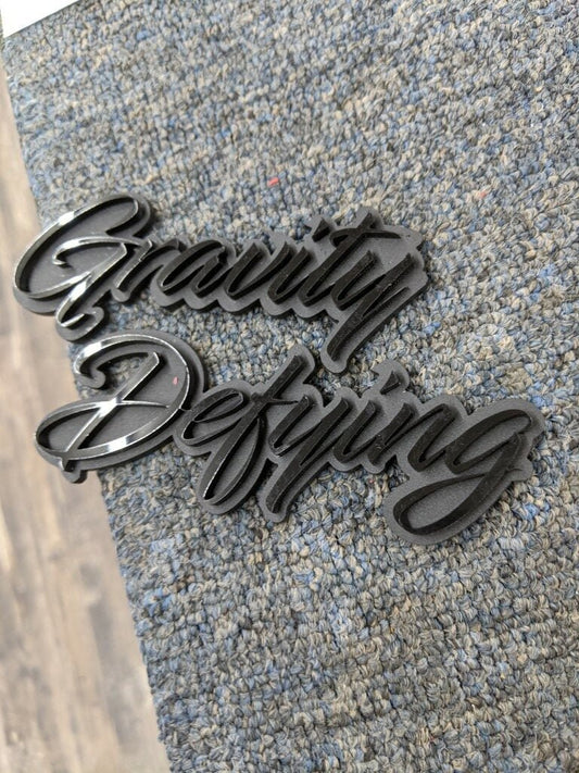 Insignia de coche Gravity Defying - Negro brillante sobre negro mate - Fuente Script