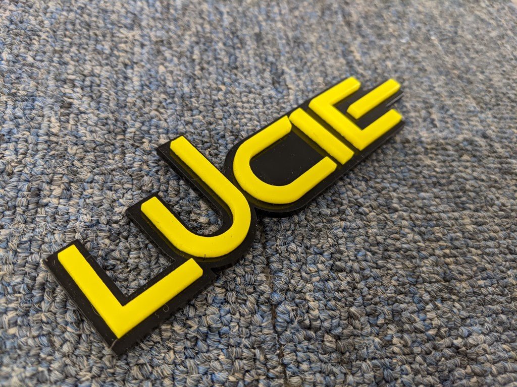 Lucie Car Badge - Yellow On Gloss Black - Squid Font - Tape Mount - Atomic Car Concepts