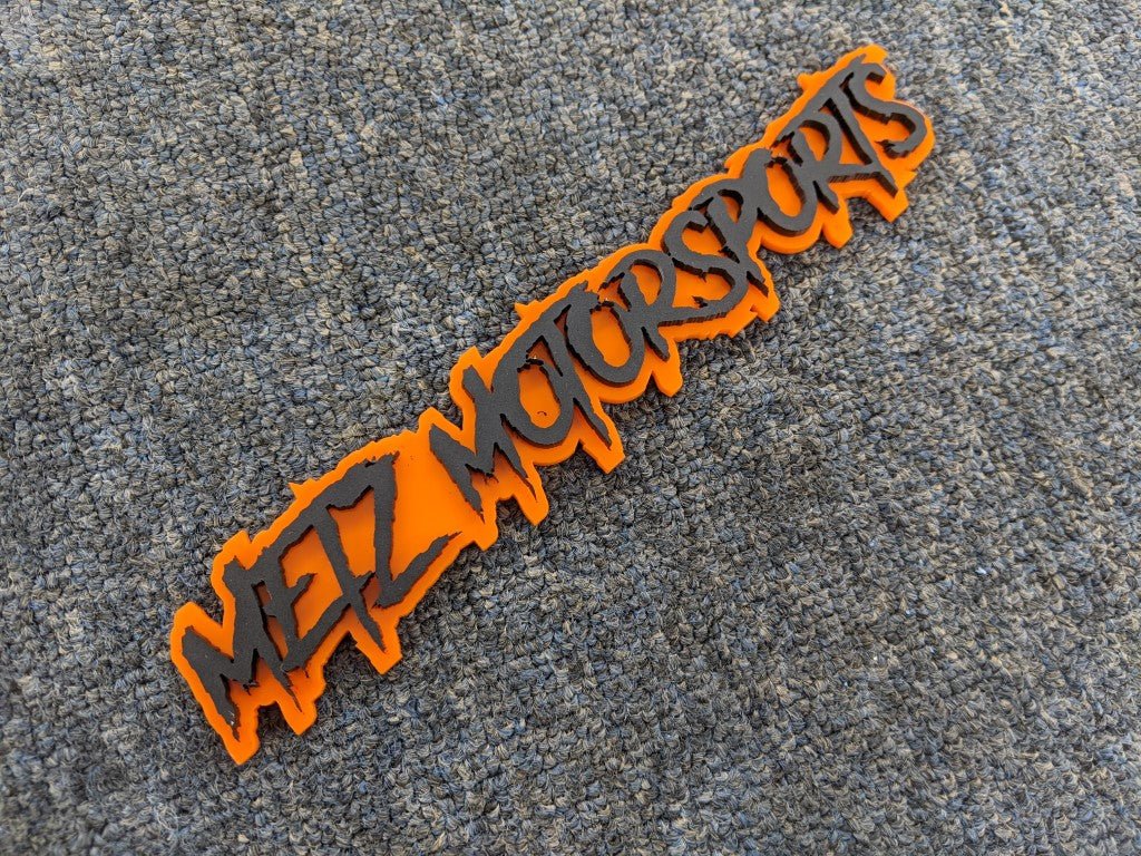 Metz Motorsports Car Badge - Matte Black On Orange - Aggressive Font - Tape Mounting - Atomic Car Concepts